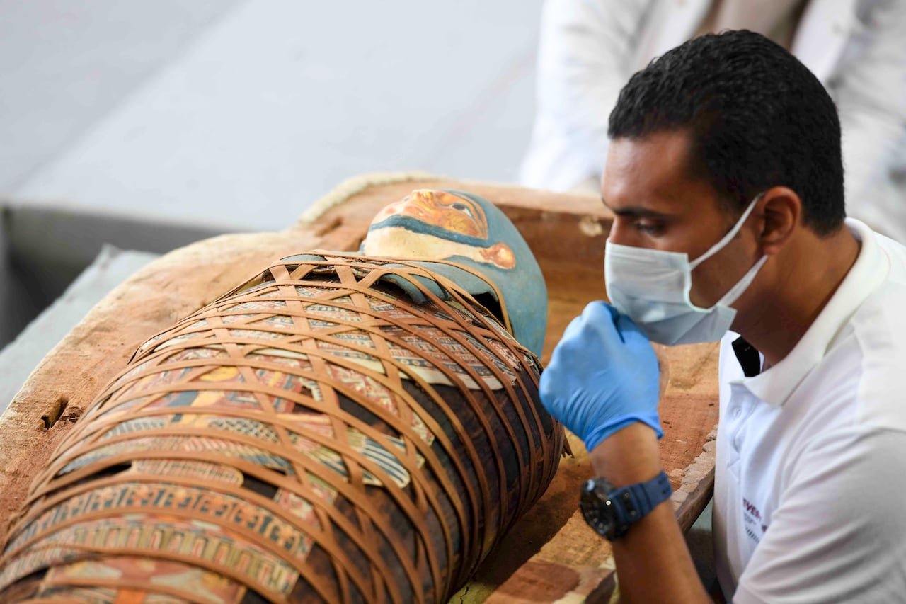 default-777 An archaeologist inspects an Egyptian mummy that is wrapped in an elaborate and colourful burial shroud. The archaeologist is wearing a medical mask and gloves.