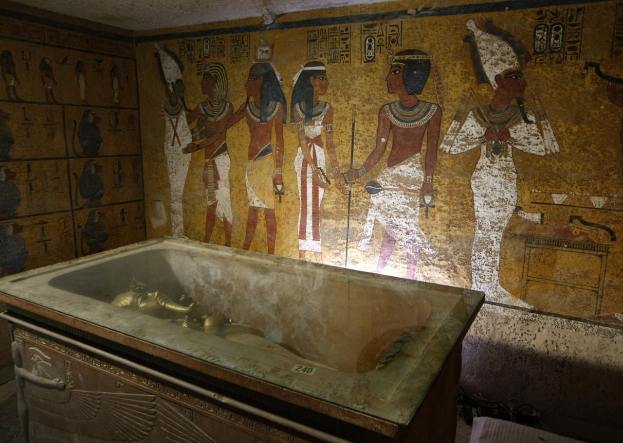 default-778 A golden sarcophagus is shown lying in an elaborate burial chamber. The wall behind it is colourfully painted with figures.